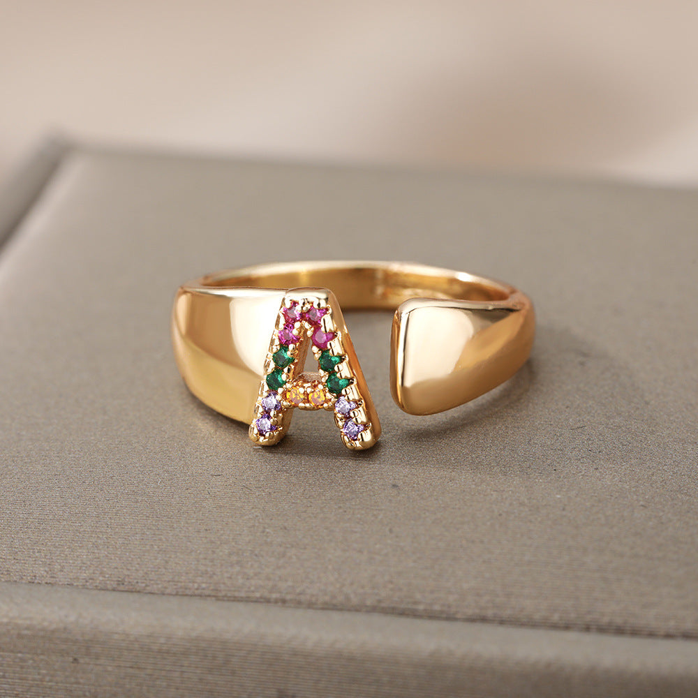 Anel com design vazado de 26 letras, cravejado com microzircônias coloridas e banhado a ouro 18K.